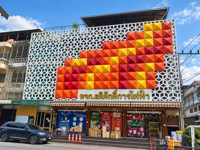 ฟาซาดร้านอดิศักดิ์การไฟฟ้า แผงตกแต่งสามมิติรูปทรงเรขาคณิตสีส้ม แดง เหลือง บนฉากตะแกรงสีขาวหน้าอาคาร