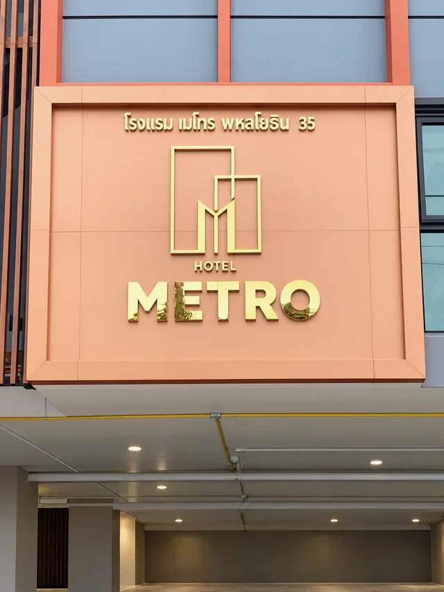 ป้าย Hotel Metro ตัวอักษรโลหะสีทองบนผนังฟาซาดสีพีช พร้อมสัญลักษณ์รูปทรงเรขาคณิต